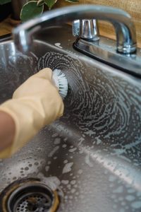 Close-up of hand with glove scrubbing a stainless steel sink with soap and brush.
